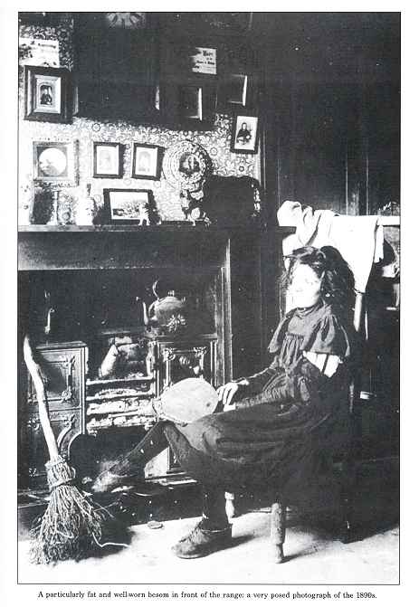 A poorly but decently clad girl sits by a fireplace holding a pair of bellows.  A besom broom leans against the wall.  Above the fireplace the mantlepiece holds china ornaments and photographs.  More framed photographs are on the wall along with a large clock.